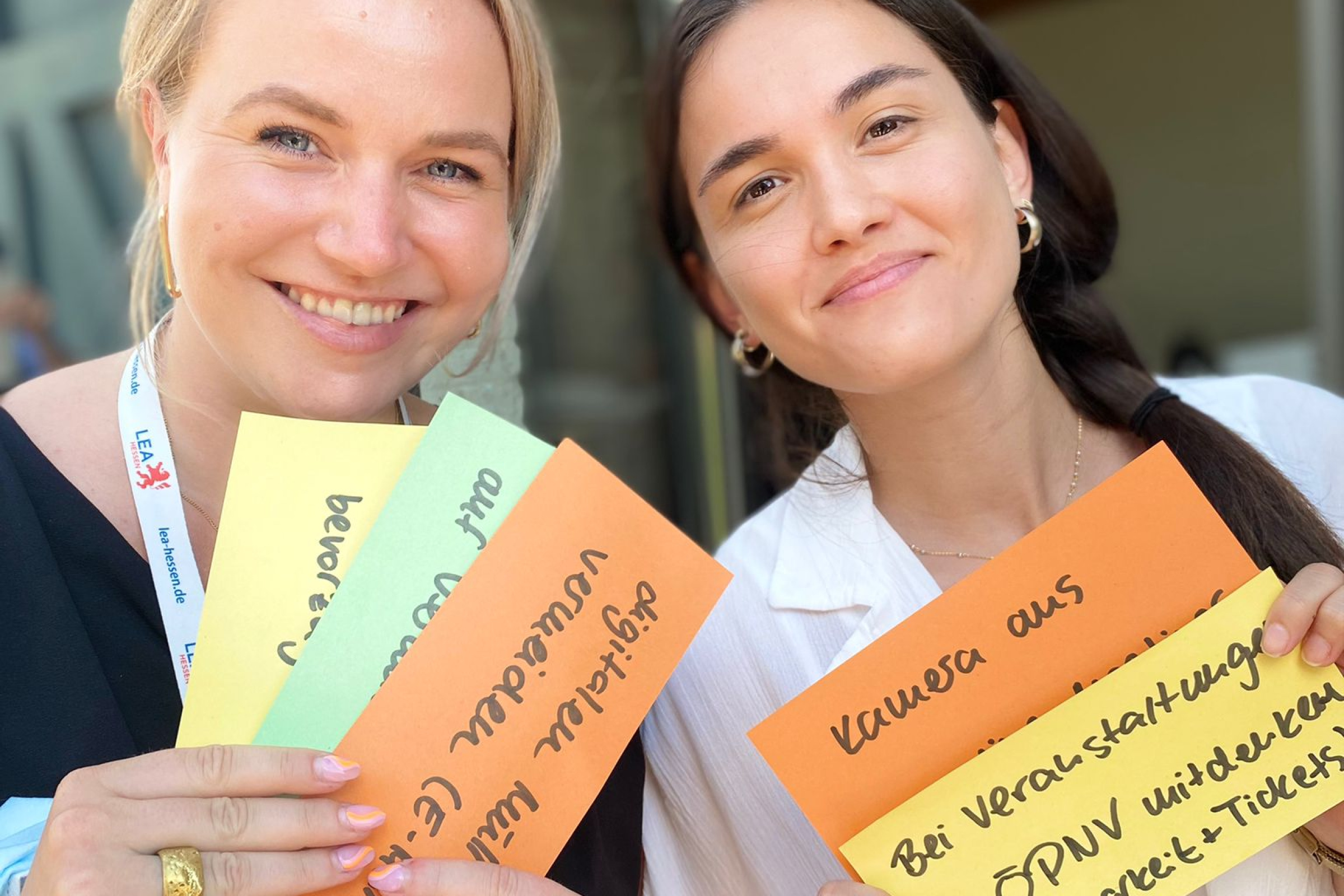 Zwei junge Frauen, eine blond, eine dunkelhaarig, stehen nebeneinander und lächeln in die Kamera. Sie halten beide mehrere farbige Kartonpapierstreifen mit Beschriftung  vor sich.